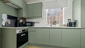 A modern kitchen with light green cabinets, a microwave, an oven, a sink, and a window with a view of a brick building. - Pear Tree - BRAND NEW Stylish City Centre Apartment - Sleeps 3 (Worcestershire)