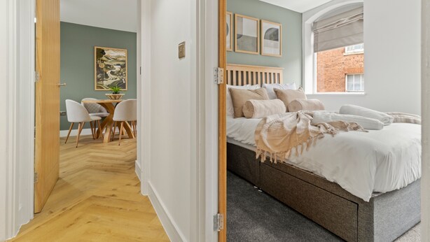 A modern bedroom with a bed, a desk with chairs, and a view of a brick building through a window. - Pear Tree - BRAND NEW Stylish City Centre Apartment - Sleeps 3 (Worcestershire)