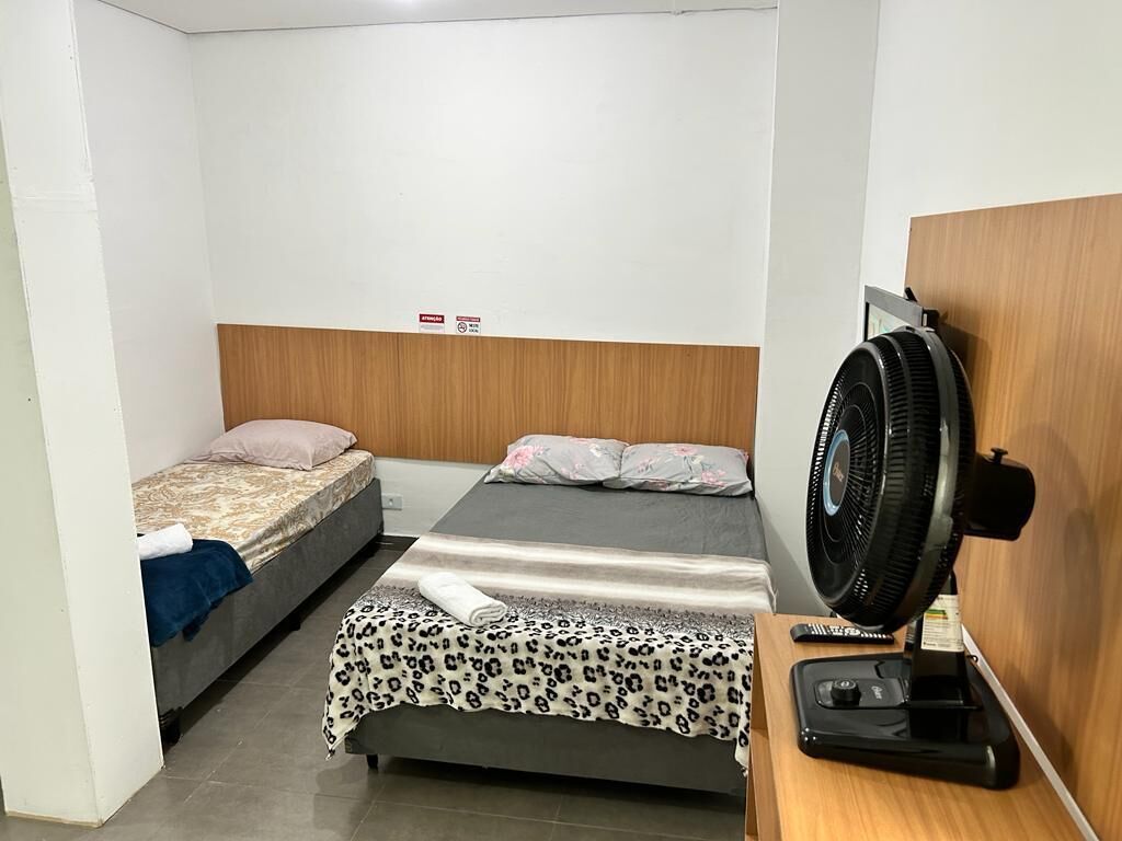A small, clean room with two single beds, a wooden headboard, a desk with a telephone, and a standing fan.