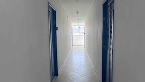 A corridor with white tiled floors and blue doors, leading to a bright room with a window. - Studio em São Paulo - Expo Center Norte (São Paulo)