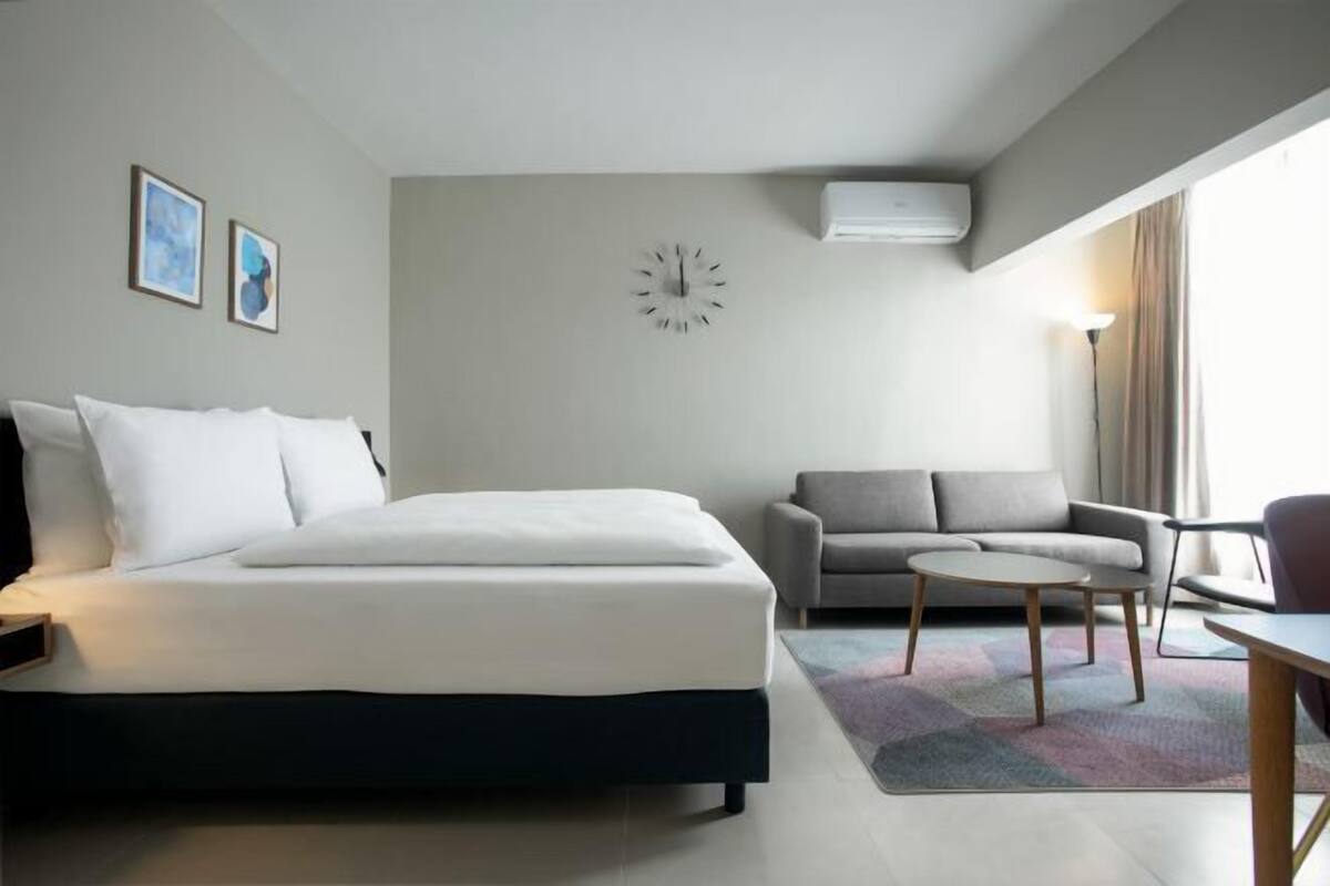 A modern hotel room with a bed, a sofa, a small table, and a clock on the wall.