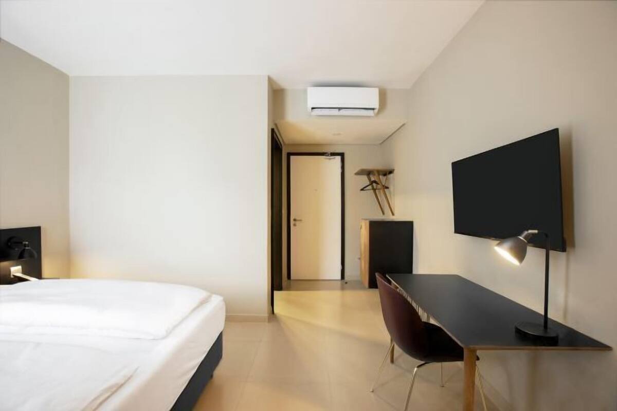 A modern hotel room with a bed, desk, chair, TV, and an air conditioning unit.
