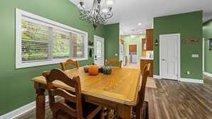 A dining room with a wooden table and chairs, a chandelier, and a view of the kitchen through a window with blinds.