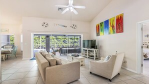 A spacious living room with a sofa, armchair, and TV stand, a balcony with a view, and a ceiling fan.