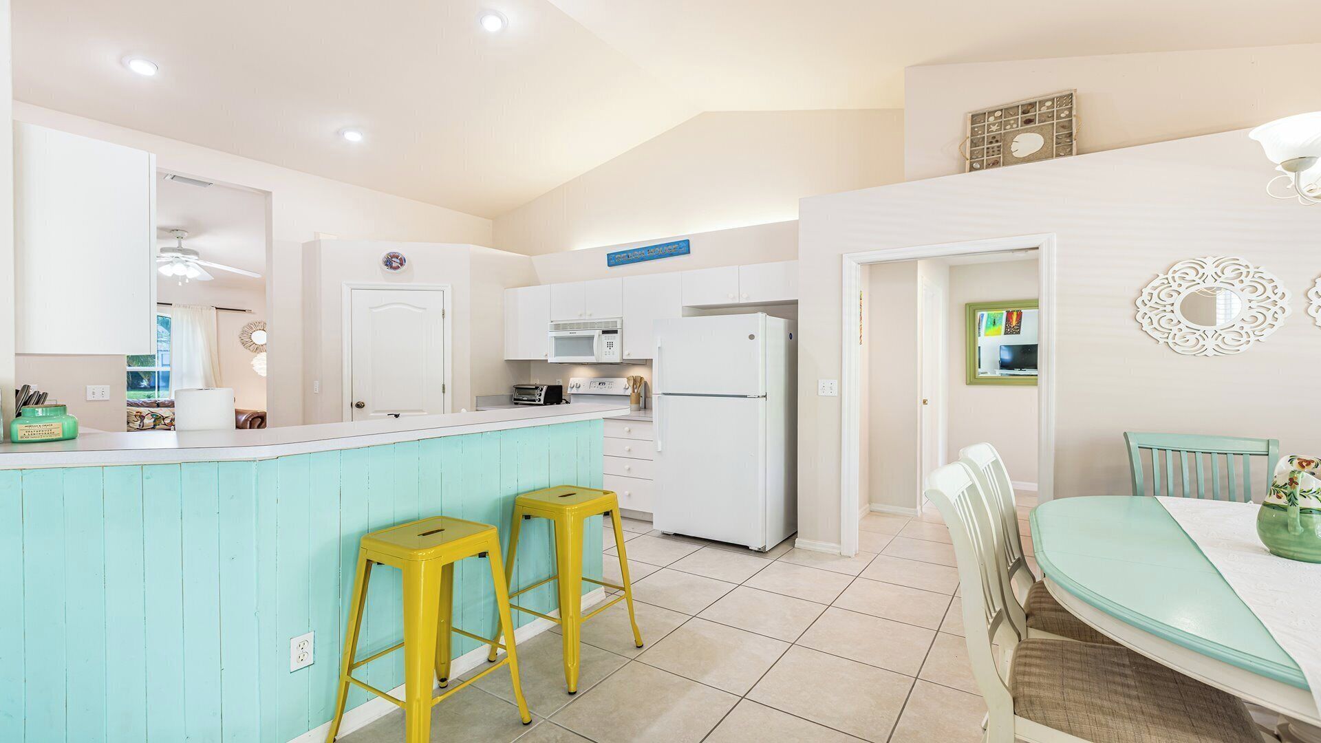 A kitchen with a turquoise island, white cabinets, and a white refrigerator. There are two yellow stools at the island and a dining table with chairs.