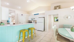 A kitchen with a turquoise island, white cabinets, and a white refrigerator. There are two yellow stools at the island and a dining table with chairs.