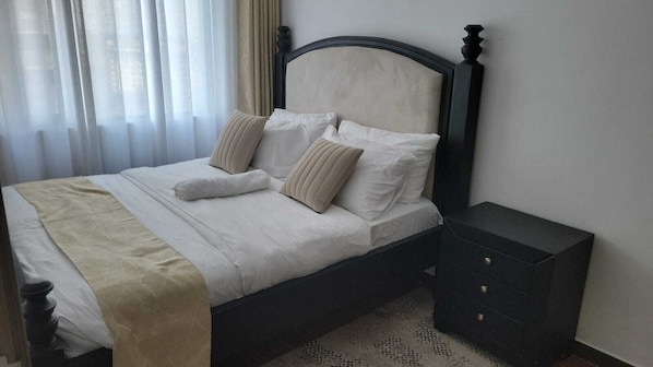 A neatly made bed with a beige bedspread and pillows, a dark wooden headboard, and a small bedside table with drawers.
