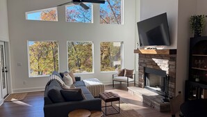 A spacious living room with a large sofa, a fireplace, a television, and multiple windows offering views of trees. - Lakefront home with Mountain Views (Mill Spring)
