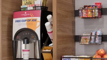 A coffee machine with a promotional sign, snack dispensers, and a fruit basket.
