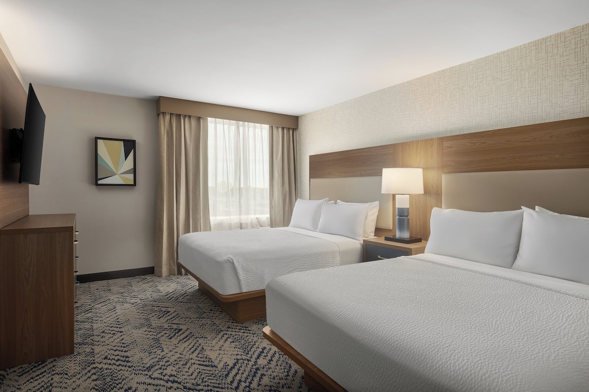 A hotel room with two beds, a wooden headboard, a flat-screen TV, a painting, and a window with curtains.