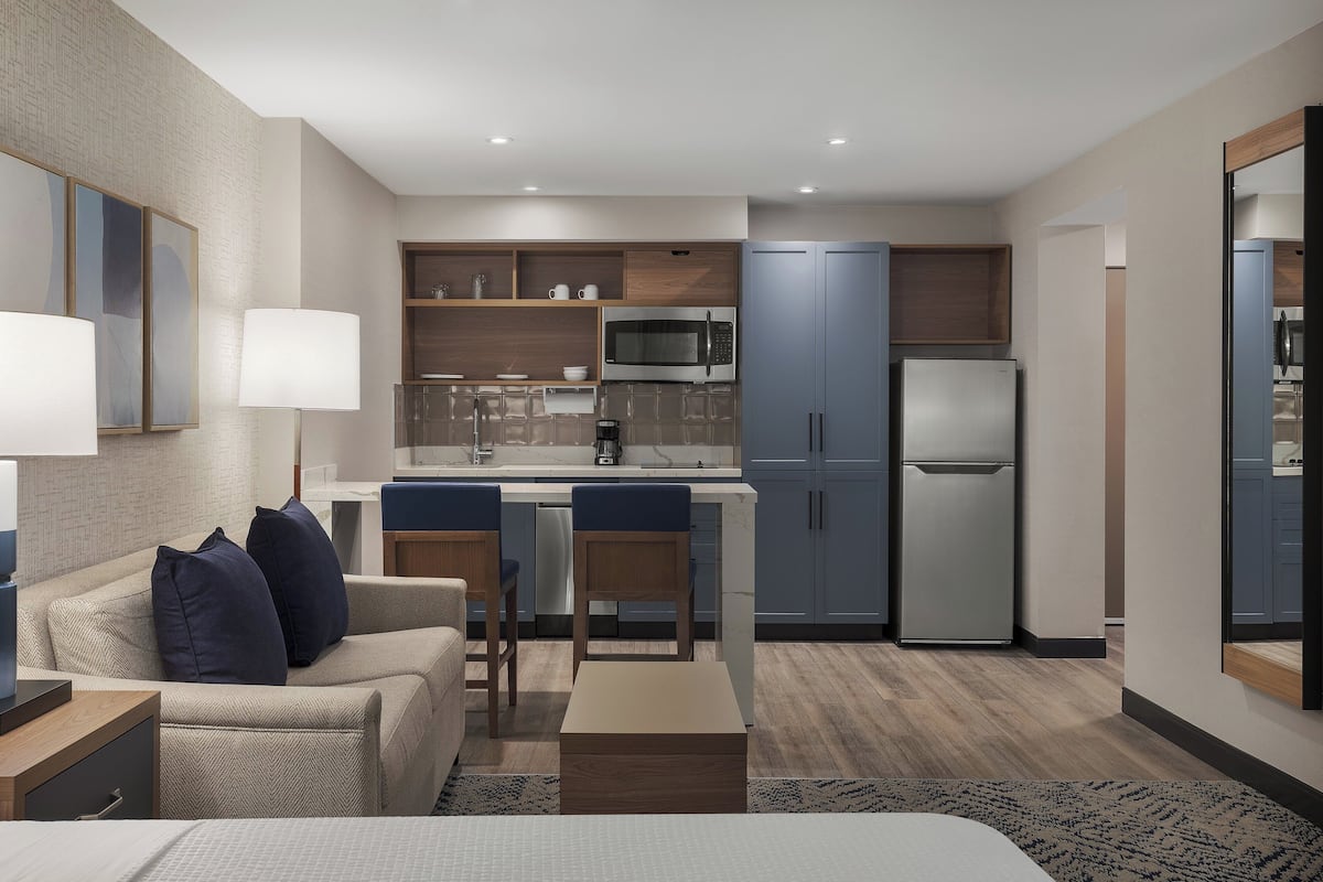 A modern hotel room with a kitchenette, a sofa, a dining table, and a bed.