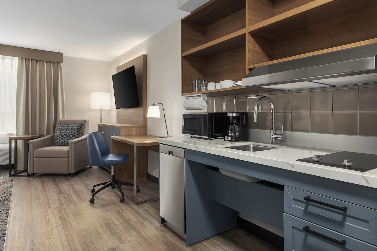 A modern hotel room with a kitchen area, a seating area with a chair and desk, and a flat-screen TV mounted on the wall.