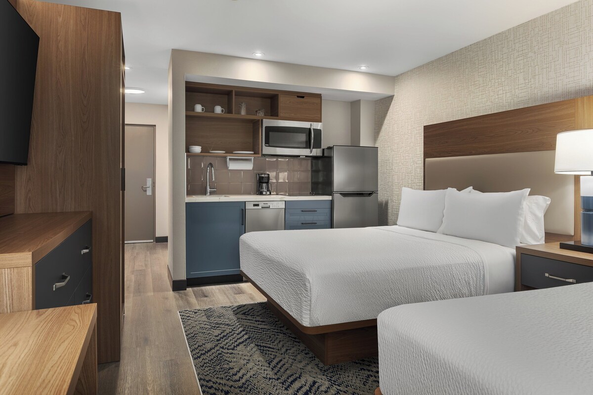 Habitación de hotel con cama, mesita de noche y una zona de cocina integrada, que incluye microondas y refrigerador.