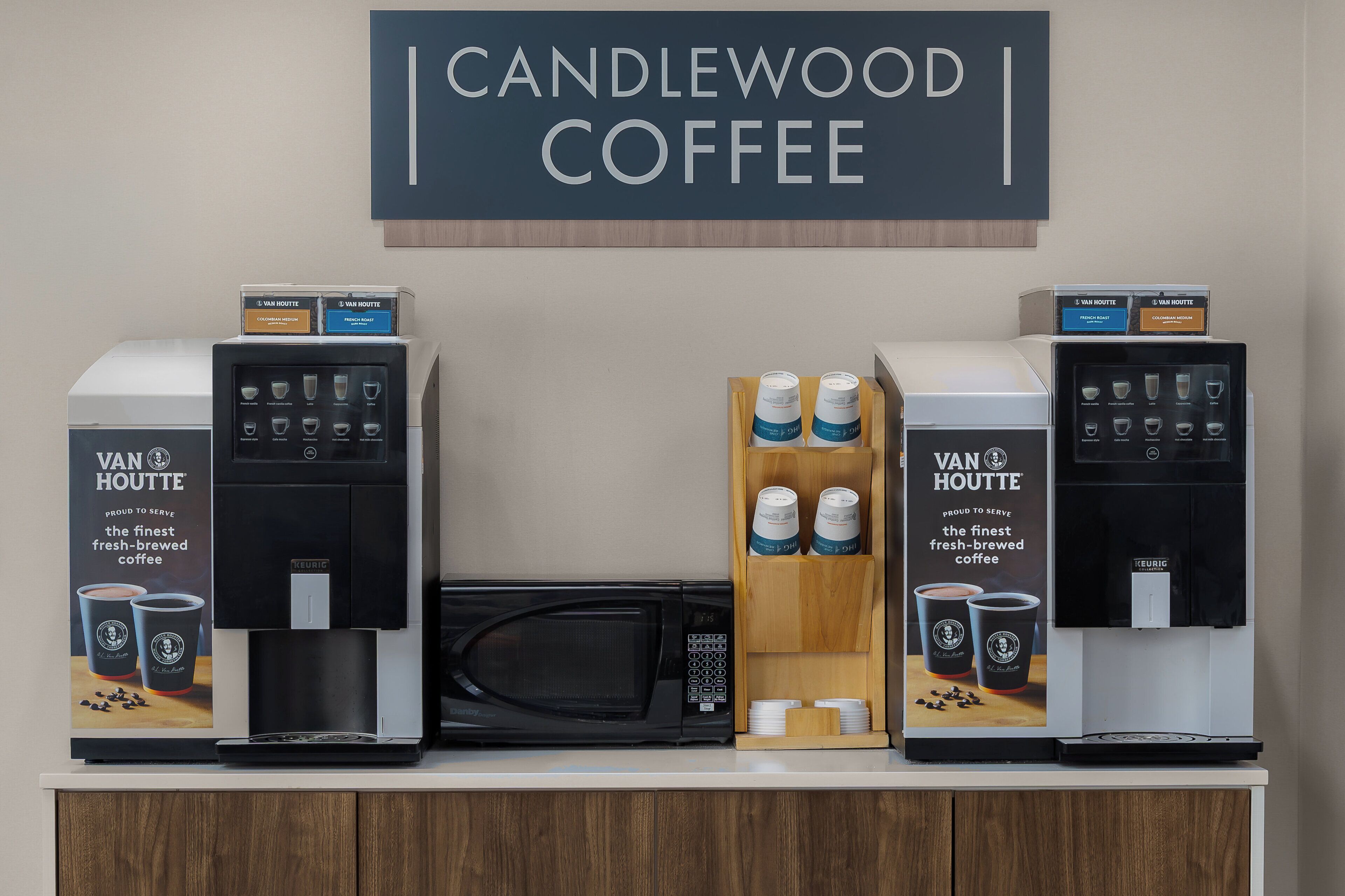 Een koffiehoek met twee koffiemachines, een bord met de tekst 'CANDLEWOOD COFFEE' en een houten vitrine met koffiekopjes.