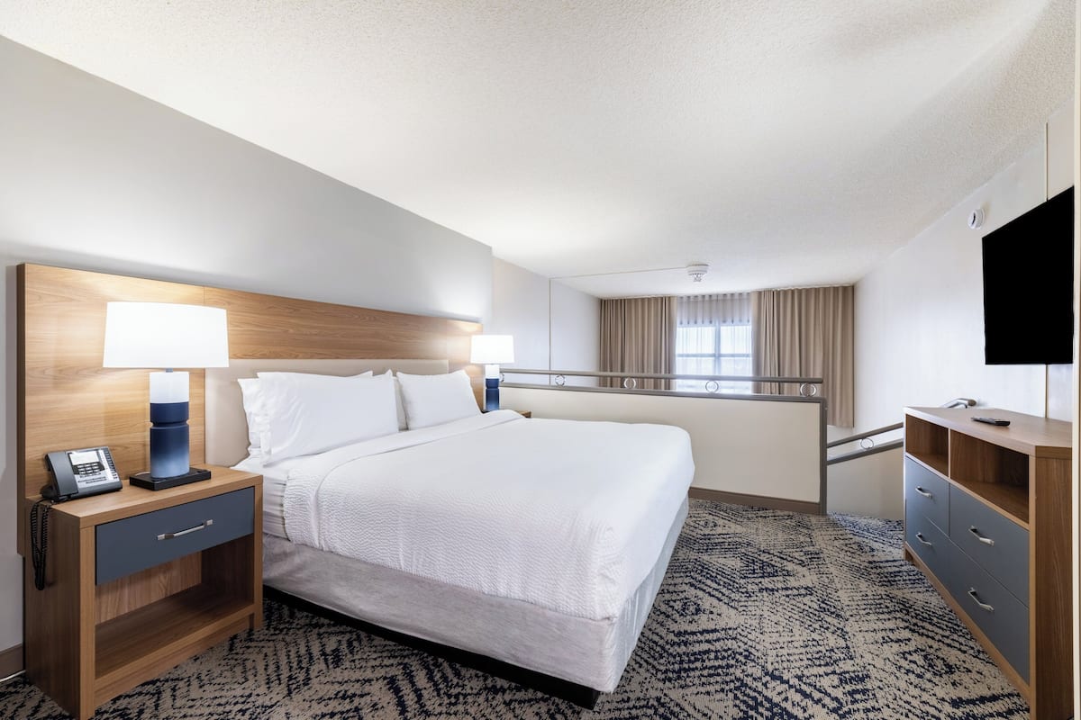 A hotel room with a bed, bedside table with a lamp, a TV, a desk with drawers, and a window with curtains.
