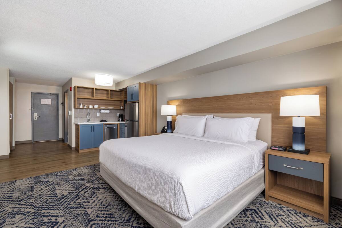 A hotel room with a large bed, bedside tables, a wooden headboard, a small kitchenette, and a patterned rug.