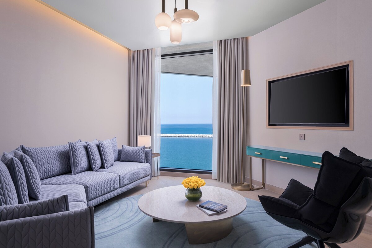 Ein modernes Hotelzimmer mit großem Fenster, einer Couch, einem Couchtisch, einem Fernseher und einem Schreibtisch.