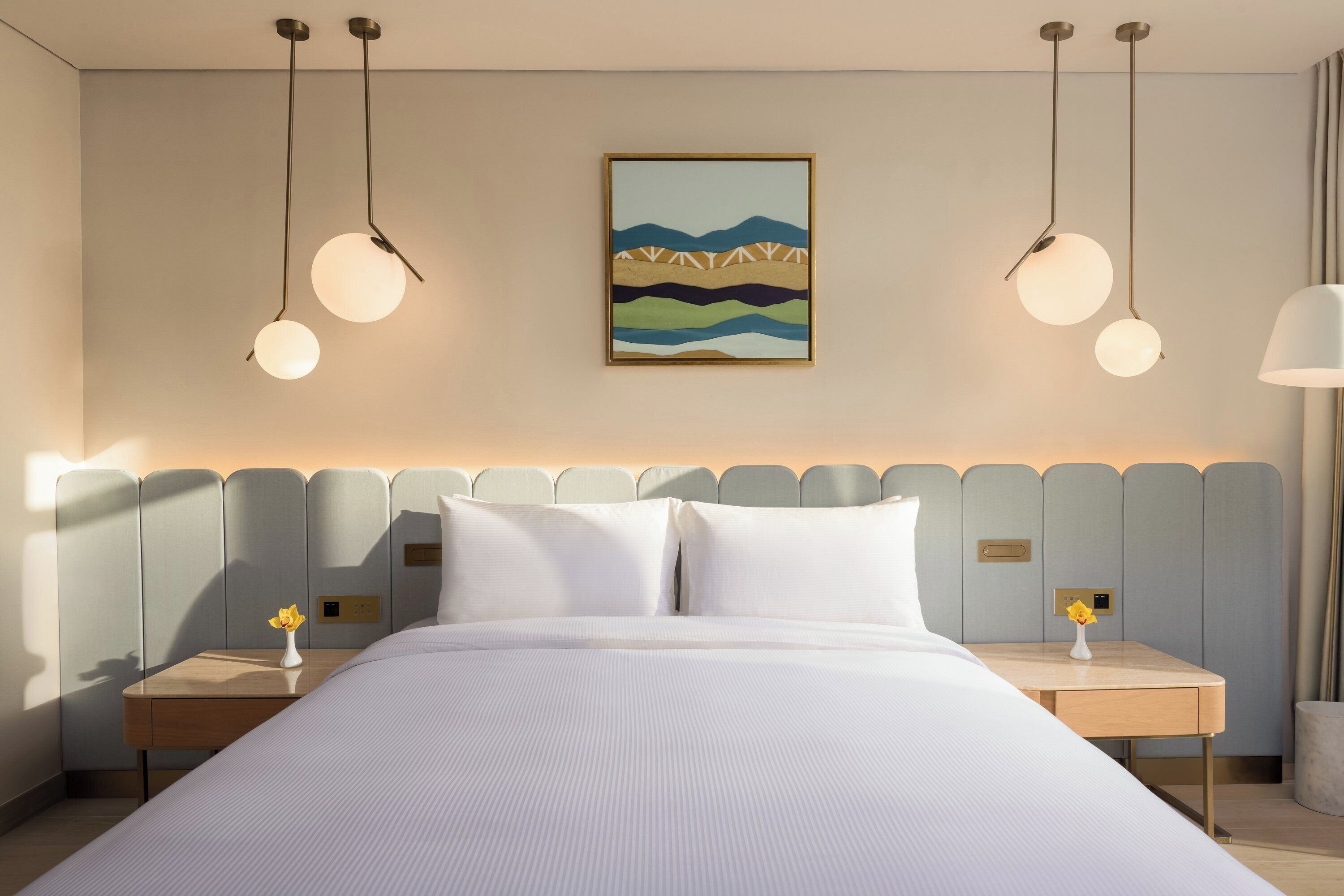 A hotel room with a large bed, two bedside tables, a wall-mounted painting, and pendant lighting.
