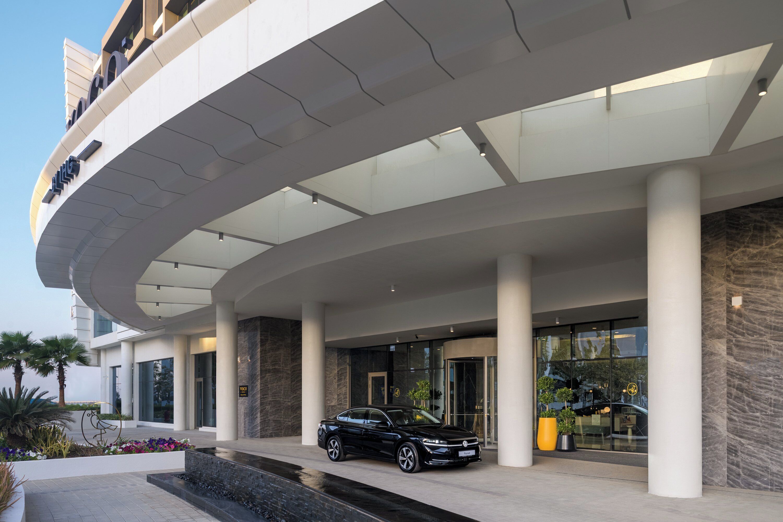 En modern byggnad med en böjd entré, en svart bil parkerad framför och en välskött trädgårdsanläggning.
