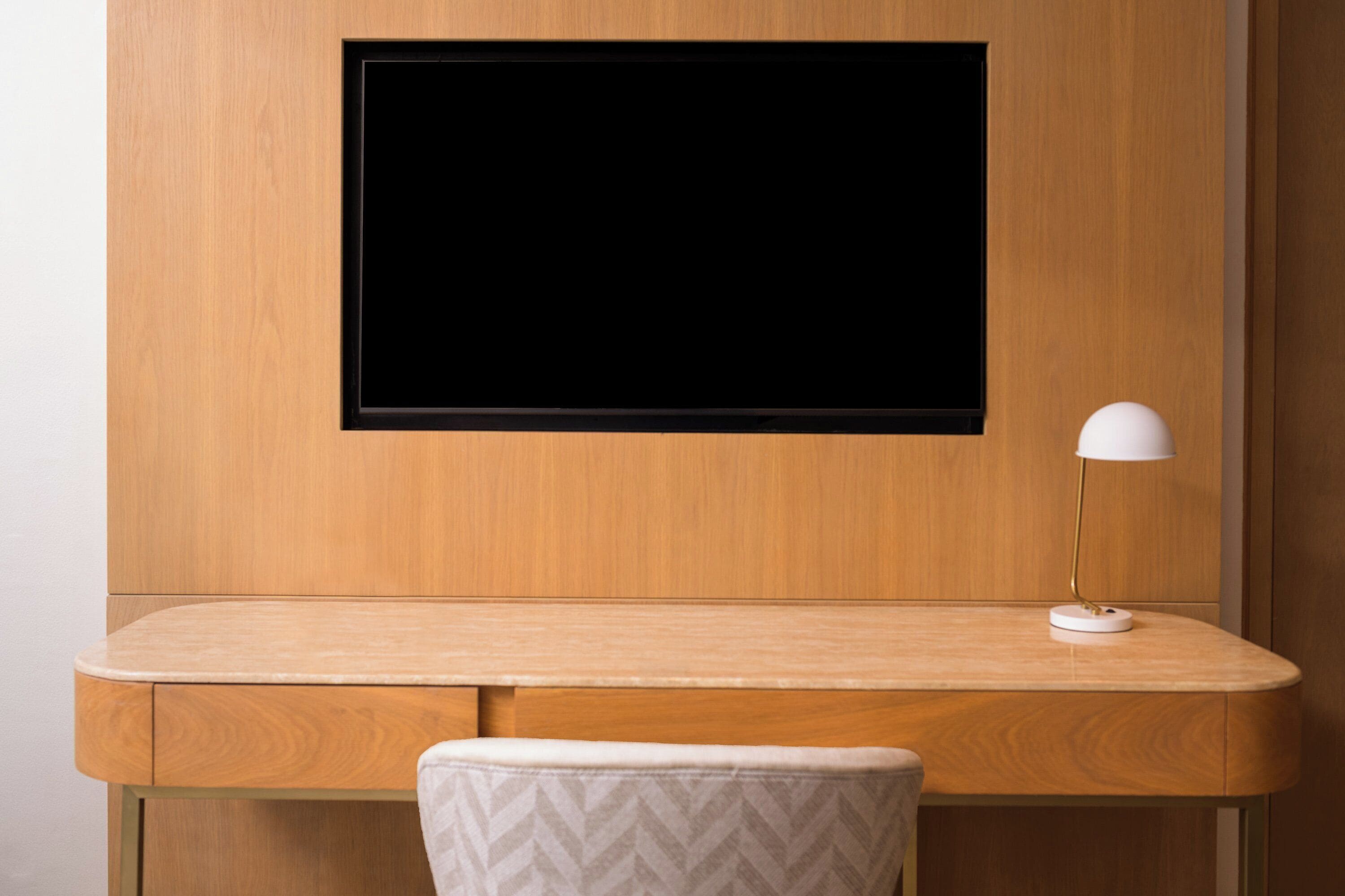 Un escritorio de madera con un televisor de pantalla plana montado encima, una lámpara de escritorio blanca y una silla acolchada.