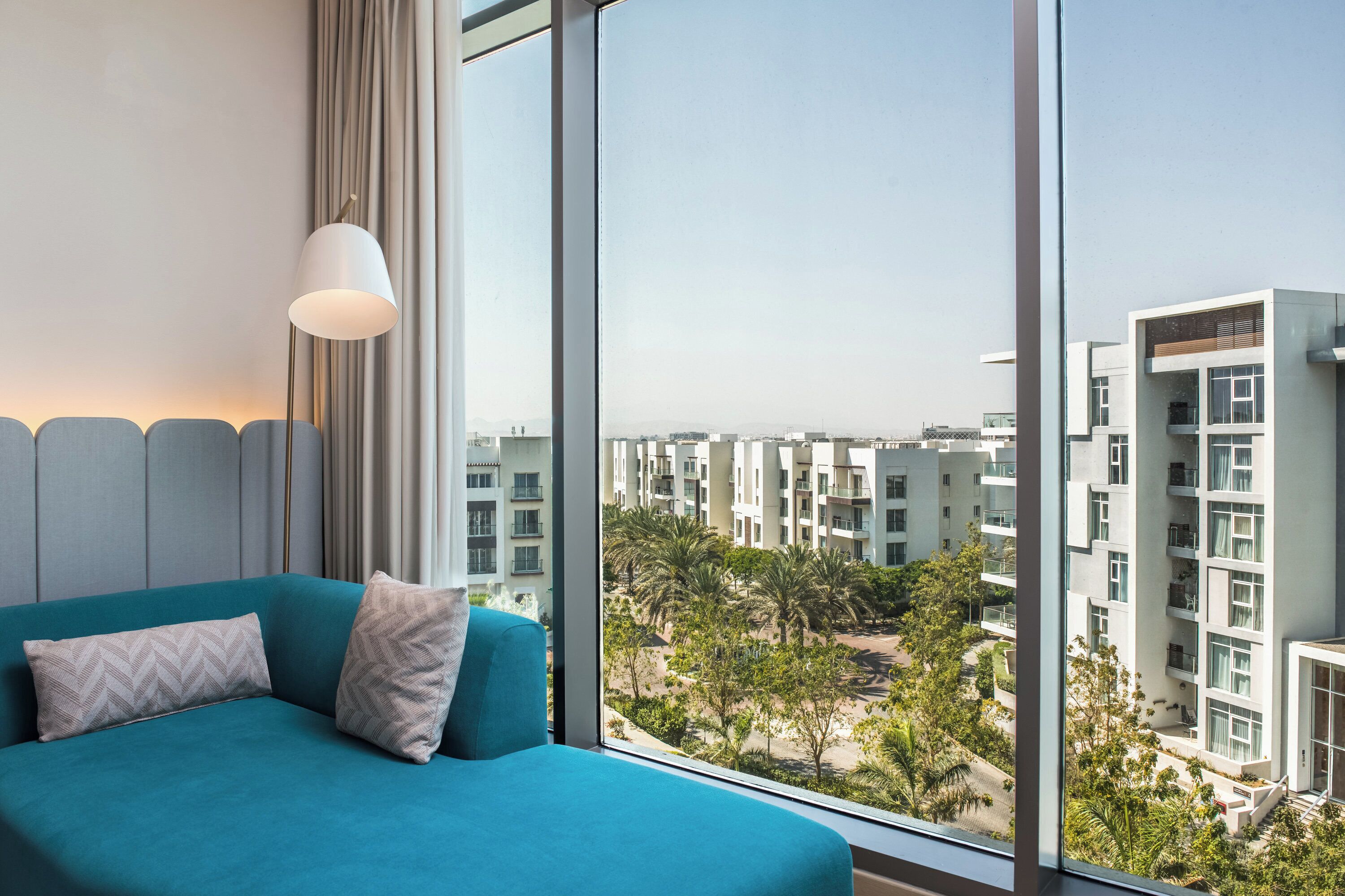A teal sofa with patterned cushions, a white lamp, and a large window with a view of modern buildings and greenery.