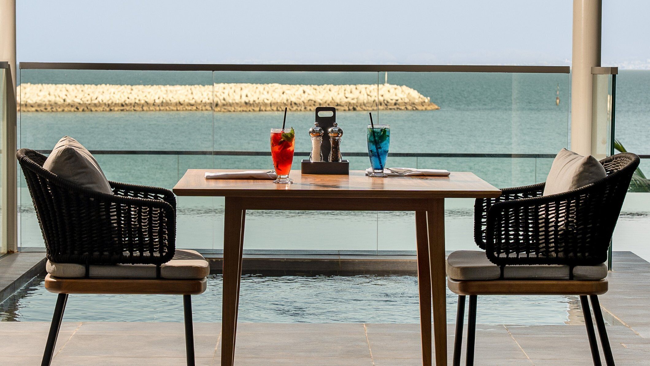 A table with two chairs, a drink, and a salt shaker on a balcony with a view of the sea.