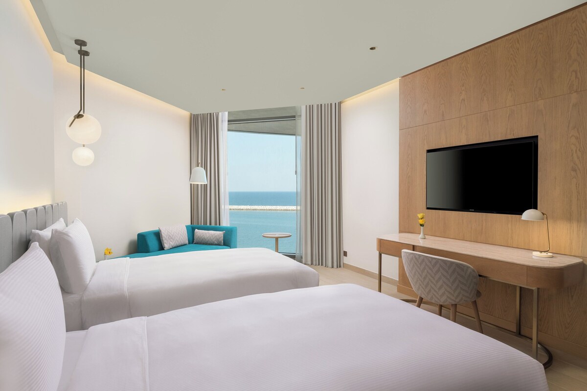 Ein Hotelzimmer mit zwei Betten, einem Schreibtisch, einem Fernseher und Meerblick.
