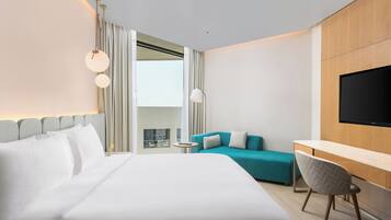A modern hotel room with a large bed, a desk, a TV, a sofa, and a view of the outdoors.