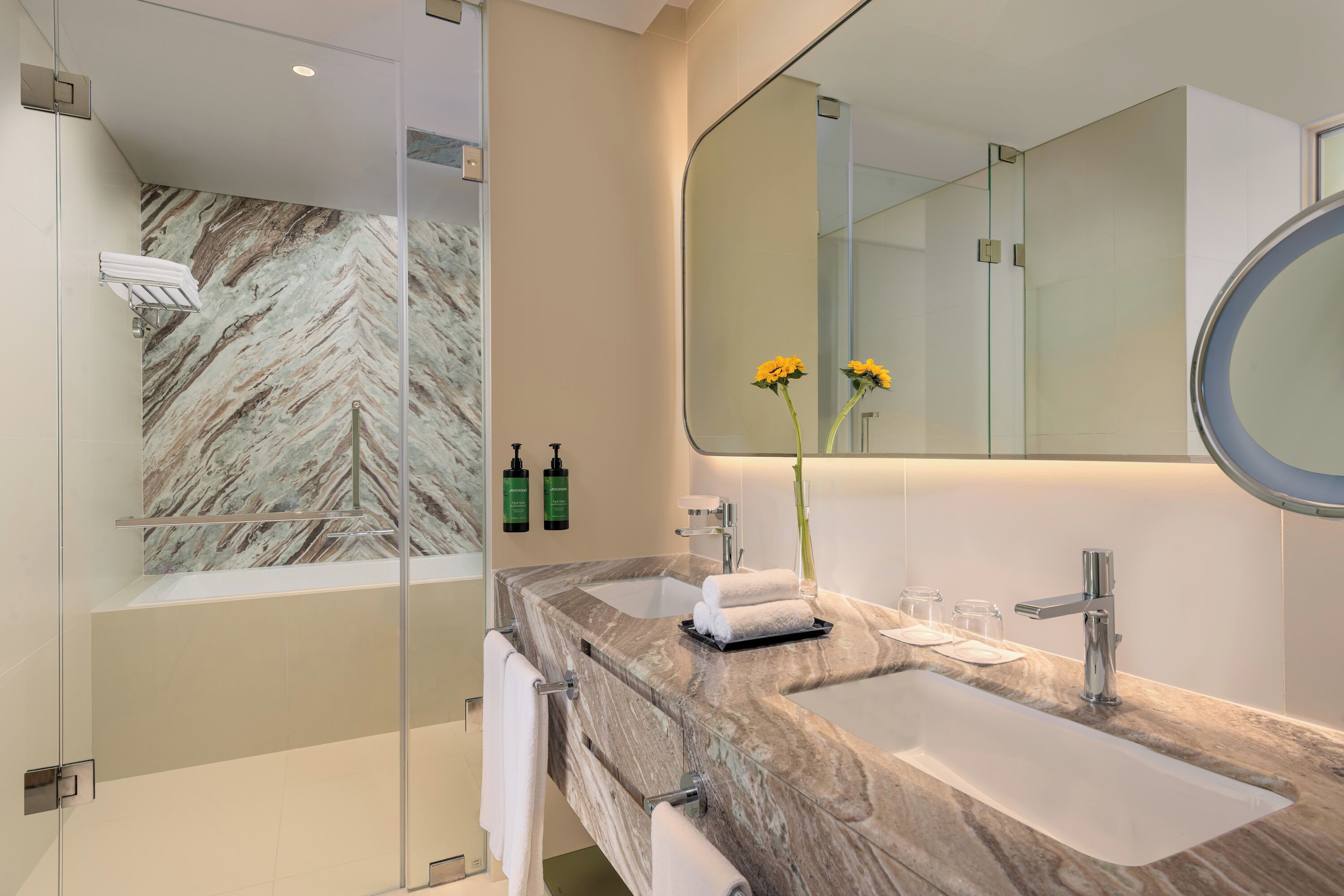 A modern bathroom with a marble wall, a large mirror, a sink, and a shower area.