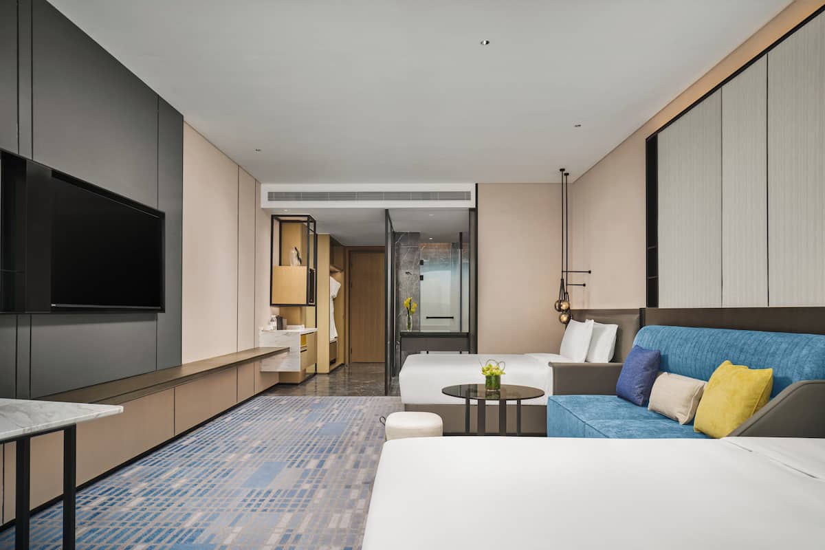 Ein modernes Hotelzimmer mit großem Flachbildfernseher, einer Couch, einem Bett, einem kleinen Tisch mit Blumenvase und einem durchgegangenen Tür sichtbaren Badezimmer.
