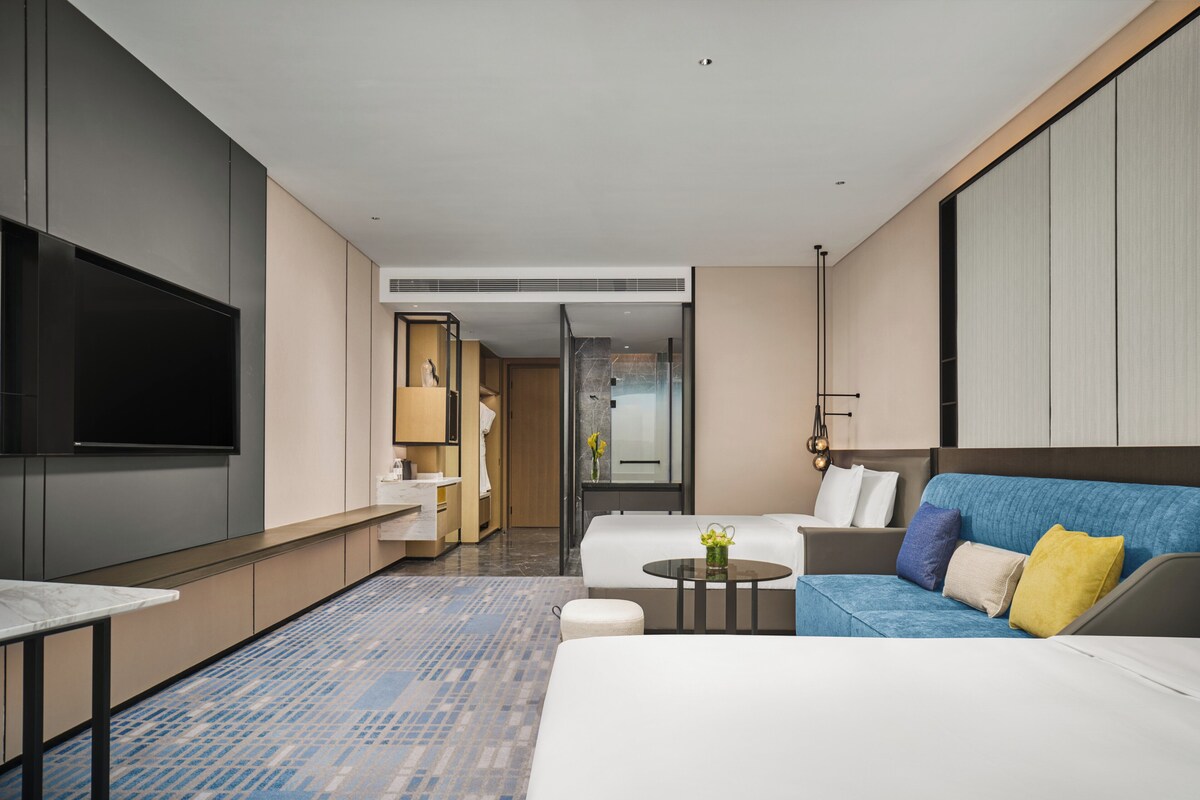 Une chambre d’hôtel moderne équipée d’un grand téléviseur à écran plat, d’un canapé, d’un lit, d’une petite table avec un vase, et d’une salle de bain visible à travers une porte ouverte.