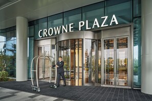 A hotel entrance with a sign reading 'CROWNE PLAZA,' a glass door entrance, and a luggage cart.