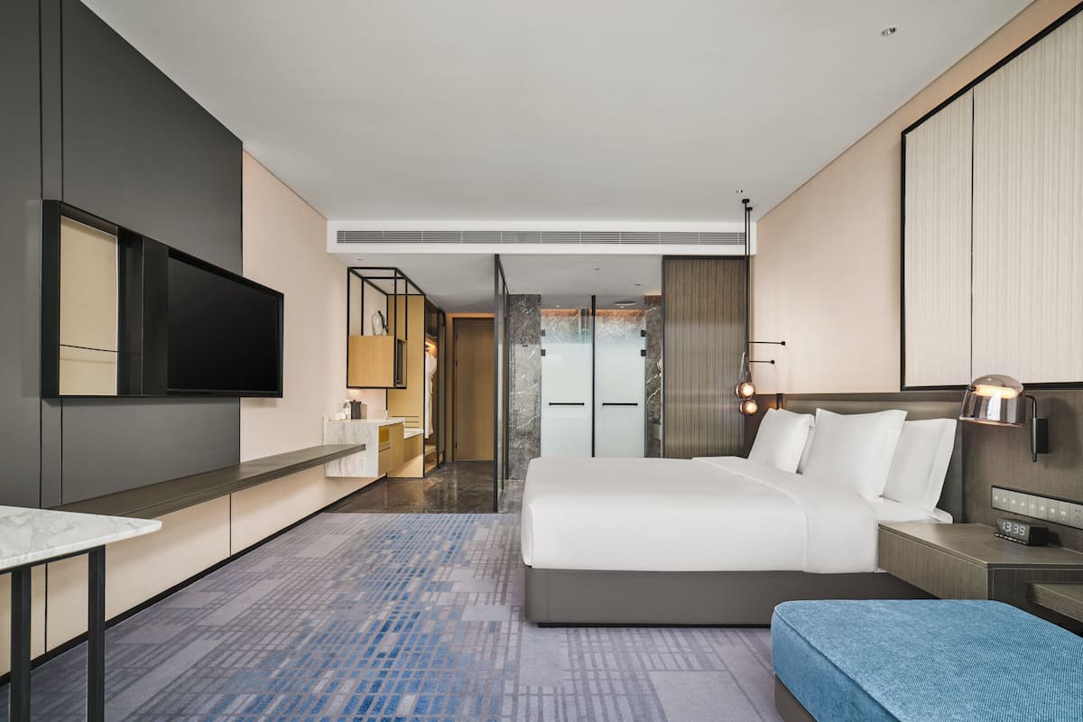 Ein modernes Hotelzimmer mit einem großen Bett, einem Flachbildfernseher, einem Schreibtisch mit Stuhl und einem durch eine Glastür einsehbaren Badezimmer.