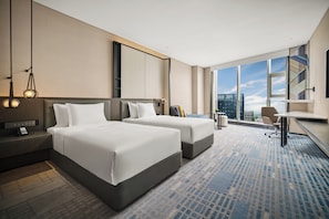 A hotel room with two beds, a desk, a chair, a large window with a city view, and a patterned carpet.
