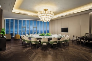A modern conference room with a large round table, multiple chairs, a chandelier, and large windows offering a view of the cityscape.