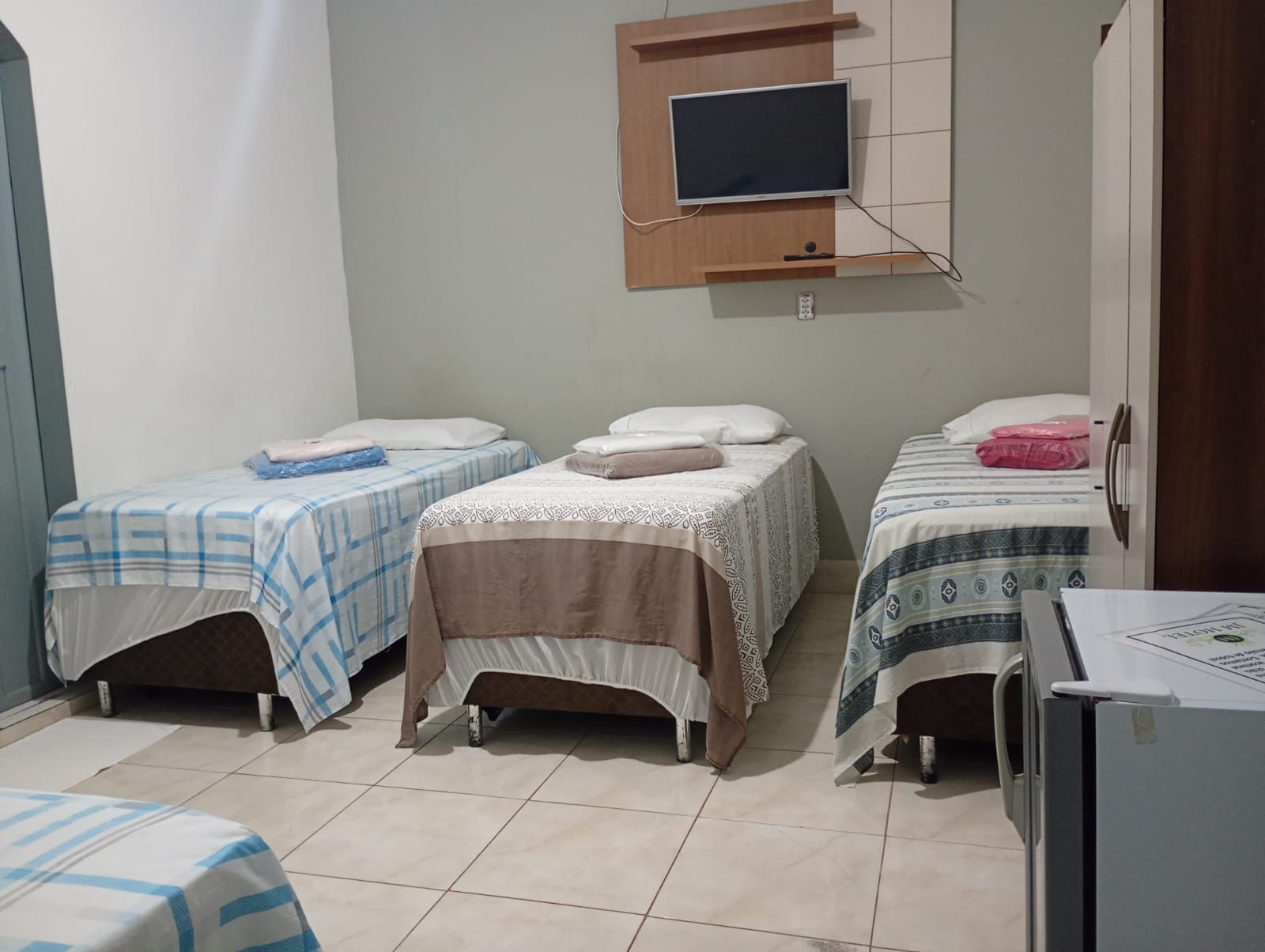 A small, tidy room with three single beds, a television mounted on the wall, and a refrigerator.