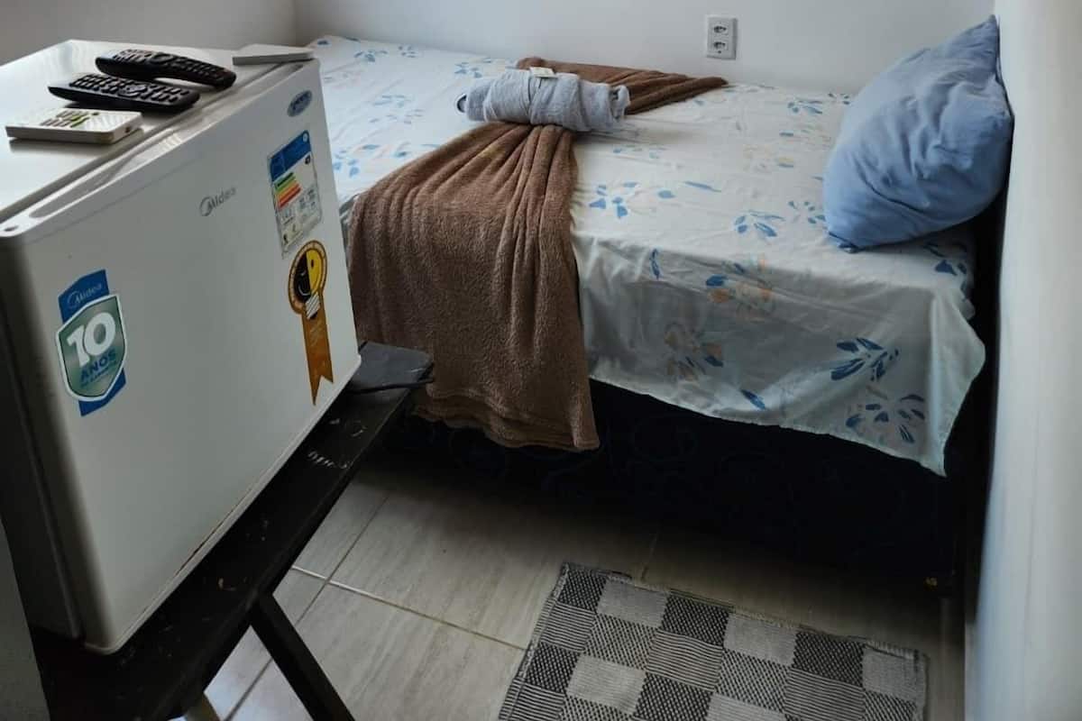Une petite pièce, de taille modeste, comprenant un lit, une petite table avec un réfrigérateur, une fenêtre avec des stores et un appareil de climatisation.