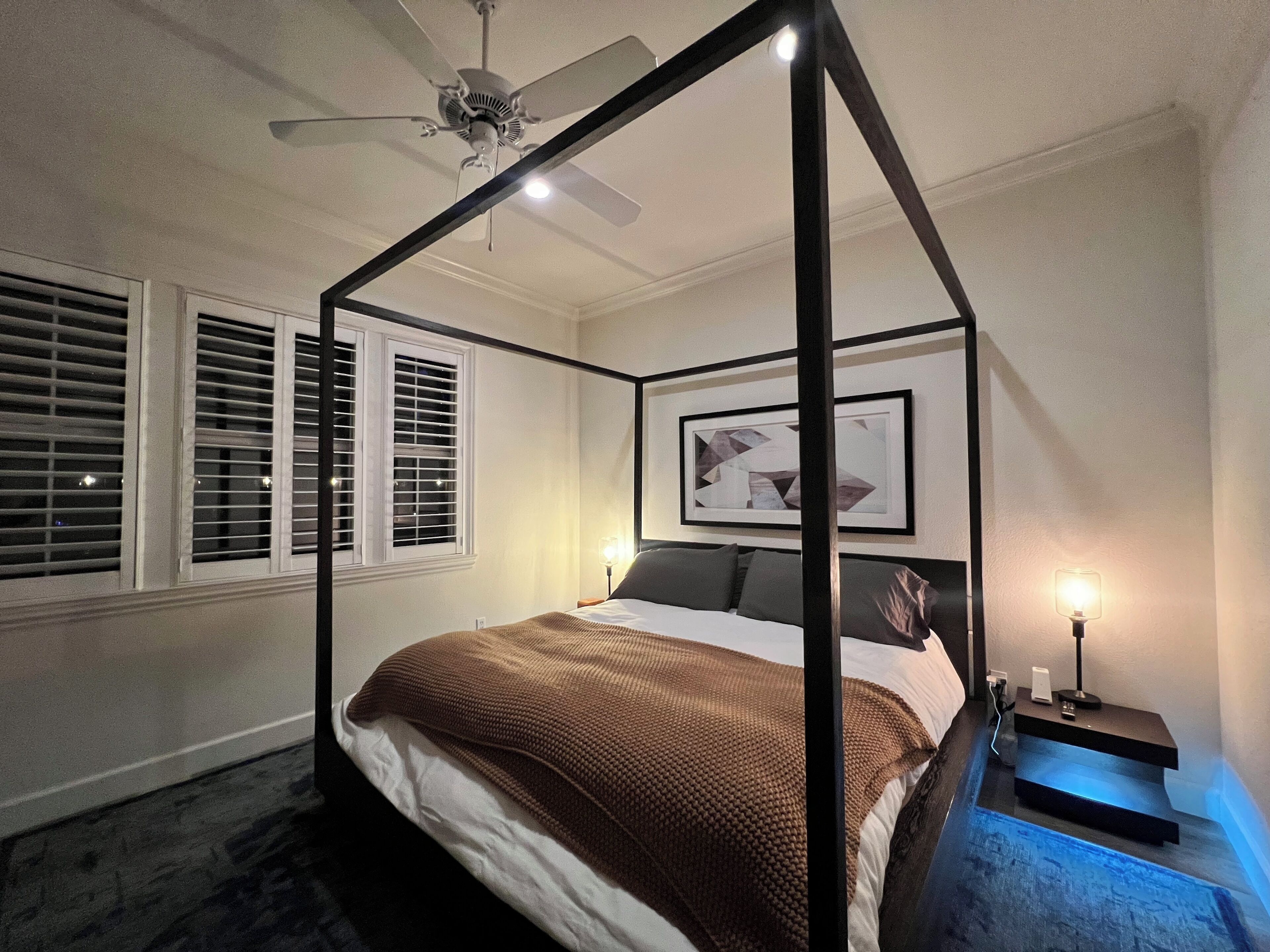 Ein Bett mit Baldachin, mit brauner Decke, ein Deckenventilator, ein gerahmtes abstraktes Gemälde und ein Nachttisch mit Lampe.