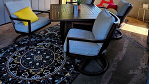 A covered patio area with a table and chairs, a ceiling fan, a dartboard, and a view of a grassy field through large windows. - “Relax, Unwind & Explore at Trailhead Hideaway – Mena’s Best Getaway!”

 (Mena)