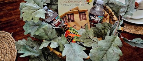 A table set with a decorative centerpiece featuring a sign for 'Too Good Gourmet Almond Tea Cookies', surrounded by leaves and a bottle of water.