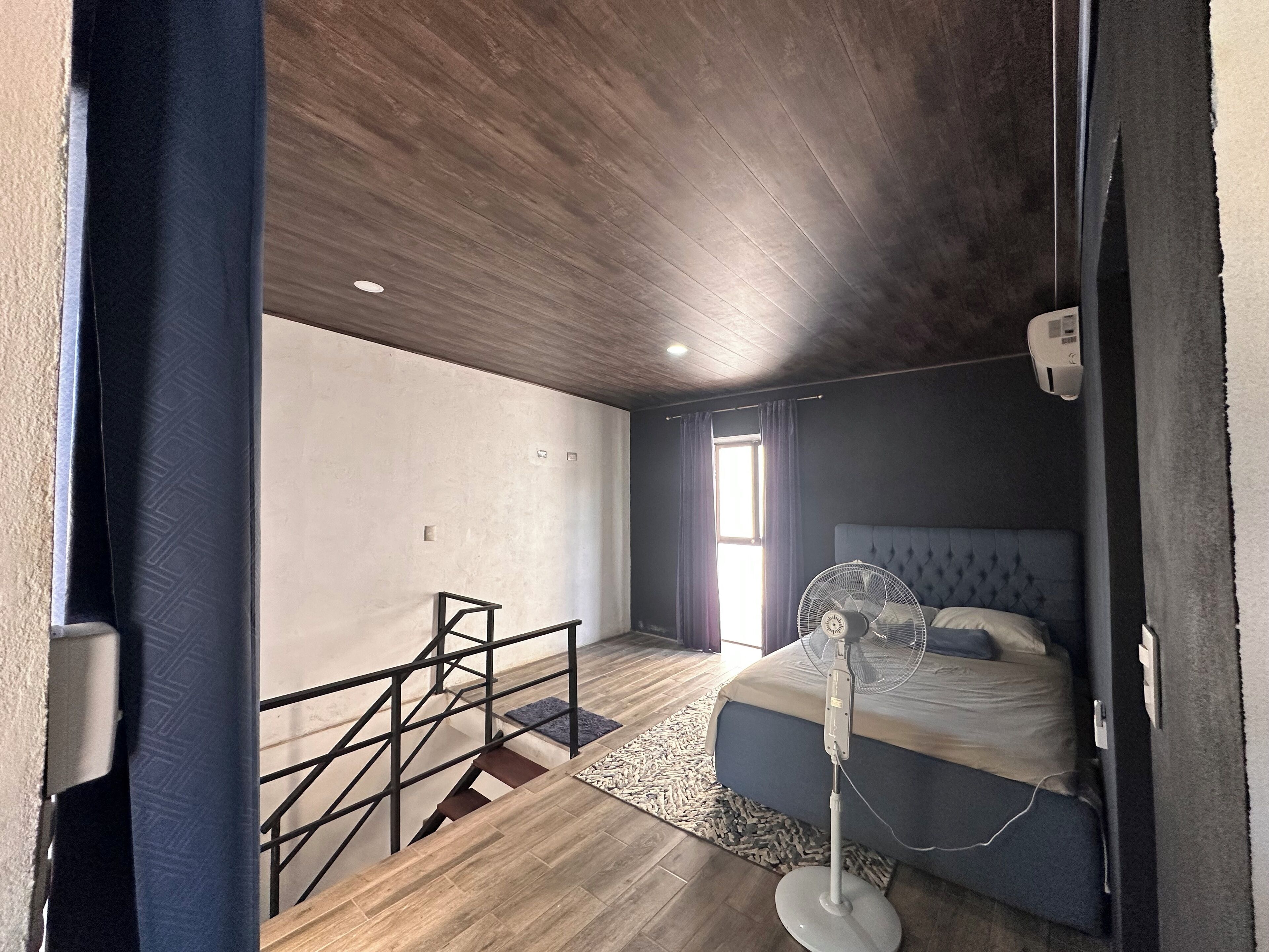 A bedroom with a bed, a standing fan, a window with curtains, a staircase, and a ceiling fan.
