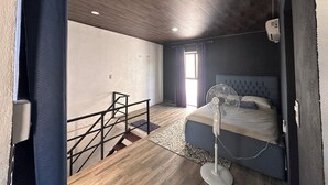 Un dormitorio con cama, ventilador, ventana con cortinas, una escalera y ventilador de techo.