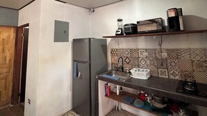 Una cocina compacta con refrigerador, microondas y estufa. Se observa un lavamanos con grifo y un estante encima que contiene una tetera y un microondas. El piso tiene baldosas estampadas, y las paredes son lisas con un pequeño enchufe eléctrico.