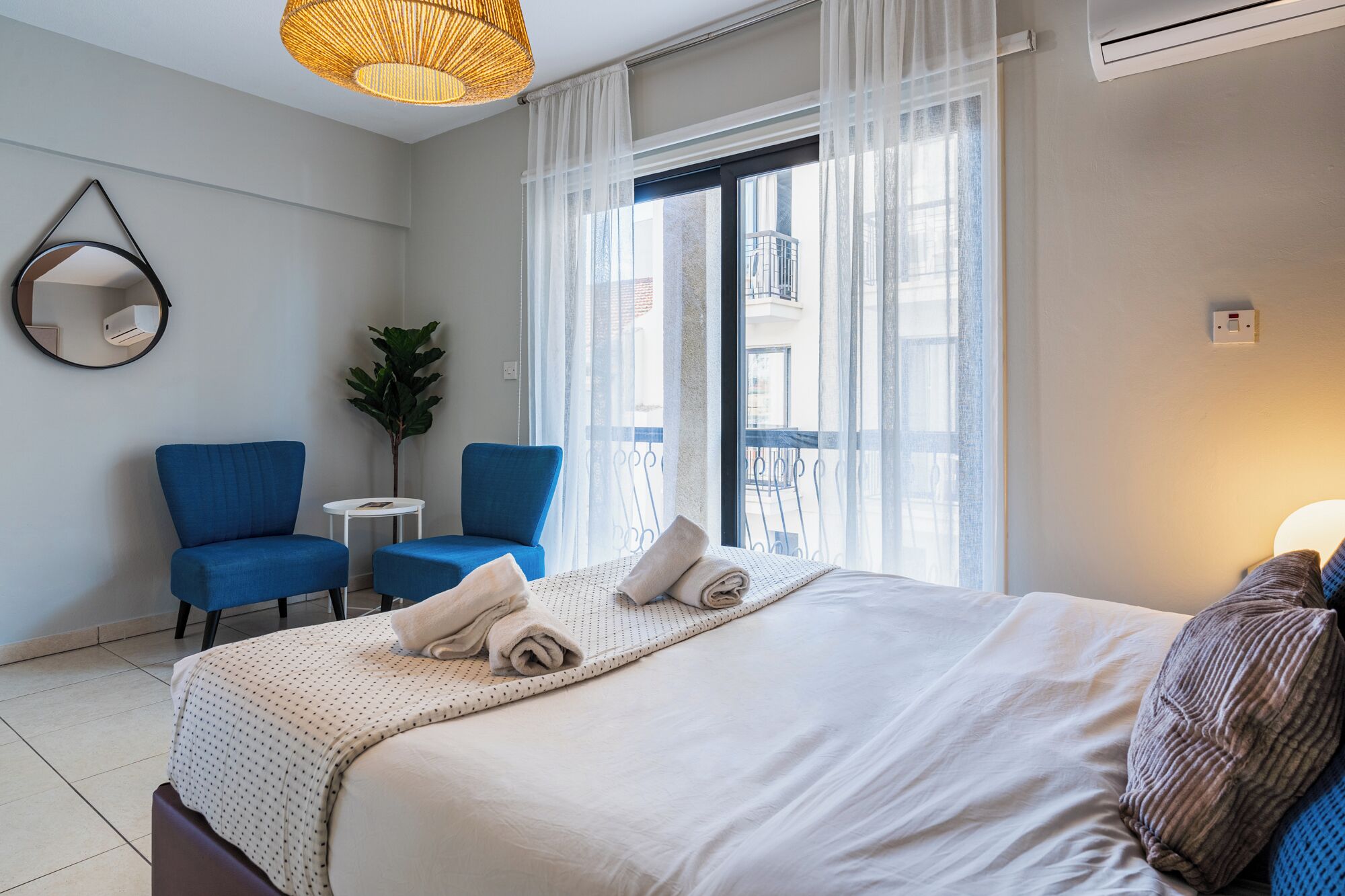 Ett hotellrum med en stor säng, två blå fåtöljer, ett litet bord och ett stort fönster med tunna, genomskinliga gardiner.