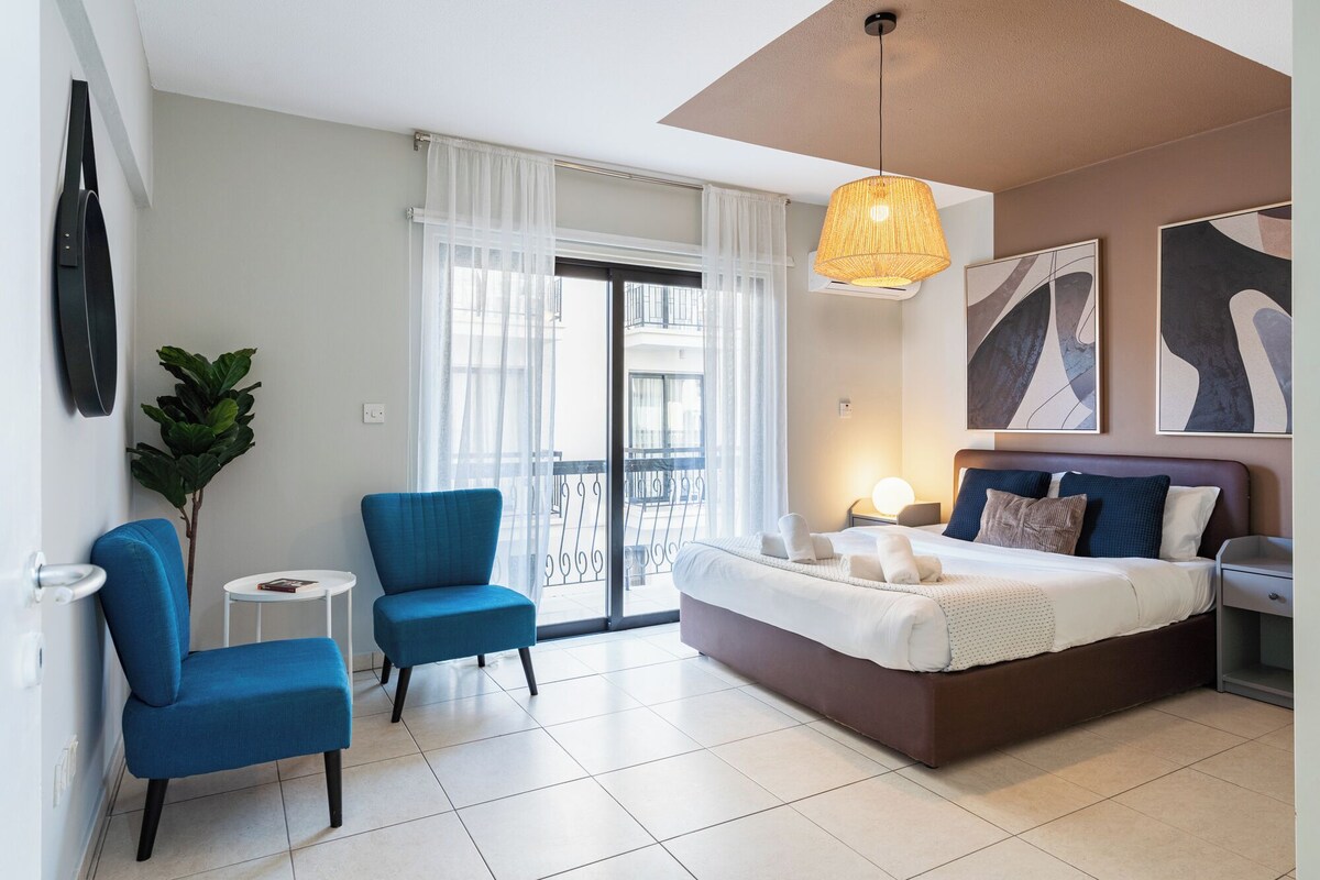 A modern hotel room with a large bed, a blue armchair, a small side table, and a balcony with a view.