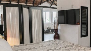 Un dormitorio con una colcha estampada, un ventanal con cortinas, un televisor de pantalla plana montado en la pared y un florero con ramas sobre un tocador.