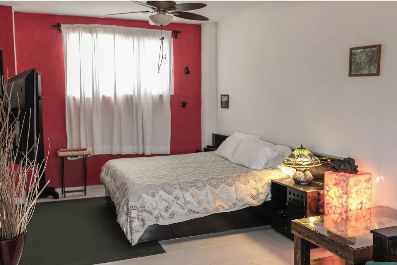 Une chambre à coucher avec un lit, une table de chevet avec une lampe, une petite table, une chaise et une fenêtre avec des rideaux.