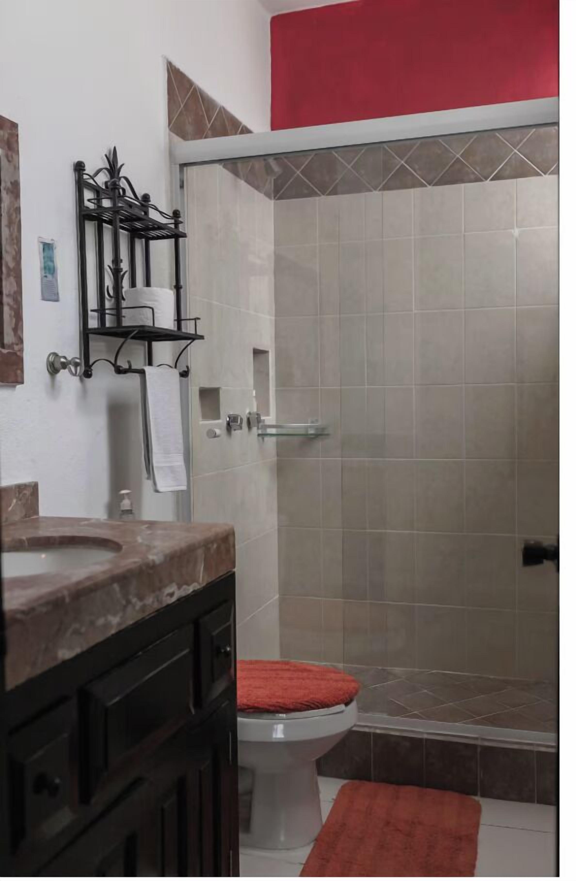 Un baño con lavamanos de mármol, inodoro con felpudo rojo y zona de ducha con paredes de azulejos.