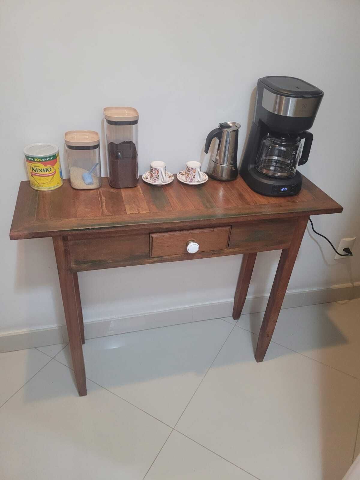 A wooden table with a drawer, a coffee maker, two cups, a thermos, and a can of Ninho.