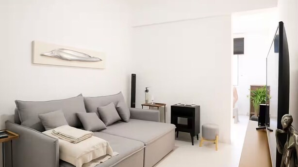 A modern living room with a grey sofa, a small side table, a black side table, and a large mirror. - 9 Andar Com Vaga de Garagem Exclusiva (Rio de Janeiro)